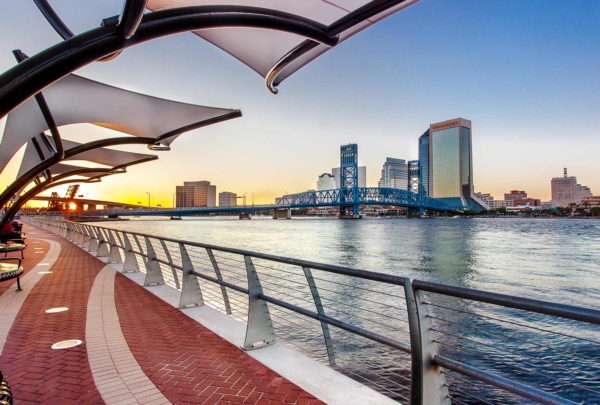 Photo of Jacksonville's Southbank Riverwalk at sunset. Red brick sidewalk alongside the river with steel guardrails. Canopies overhang the sidewalks. Jacksonville skyline in background.