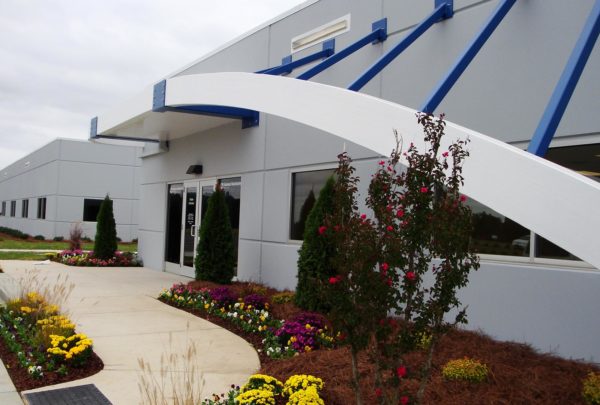 Front entrance of Turbomeca TMM Manufacturing Facility. Awning supported by blue structures. Floral landscaping.